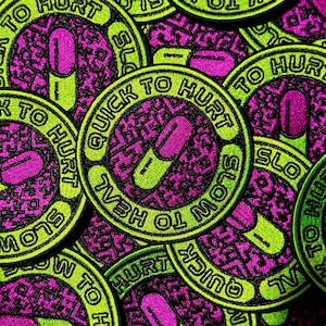 May include: A circular embroidered patch featuring a neon green border with the black text QUICK TO HURT SLOW TO HEAL. The center has a magenta and neon green pill icon set against a black and magenta patterned background with a stitched texture.