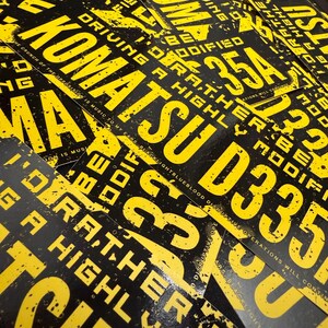 Puede incluir: Una colecci&oacute;n de impresiones gr&aacute;ficas en negro y amarillo. Las impresiones presentan las palabras "KOMATSU D335A" en grandes letras amarillas, junto con otro texto. El dise&ntilde;o general tiene una est&eacute;tica grunge.