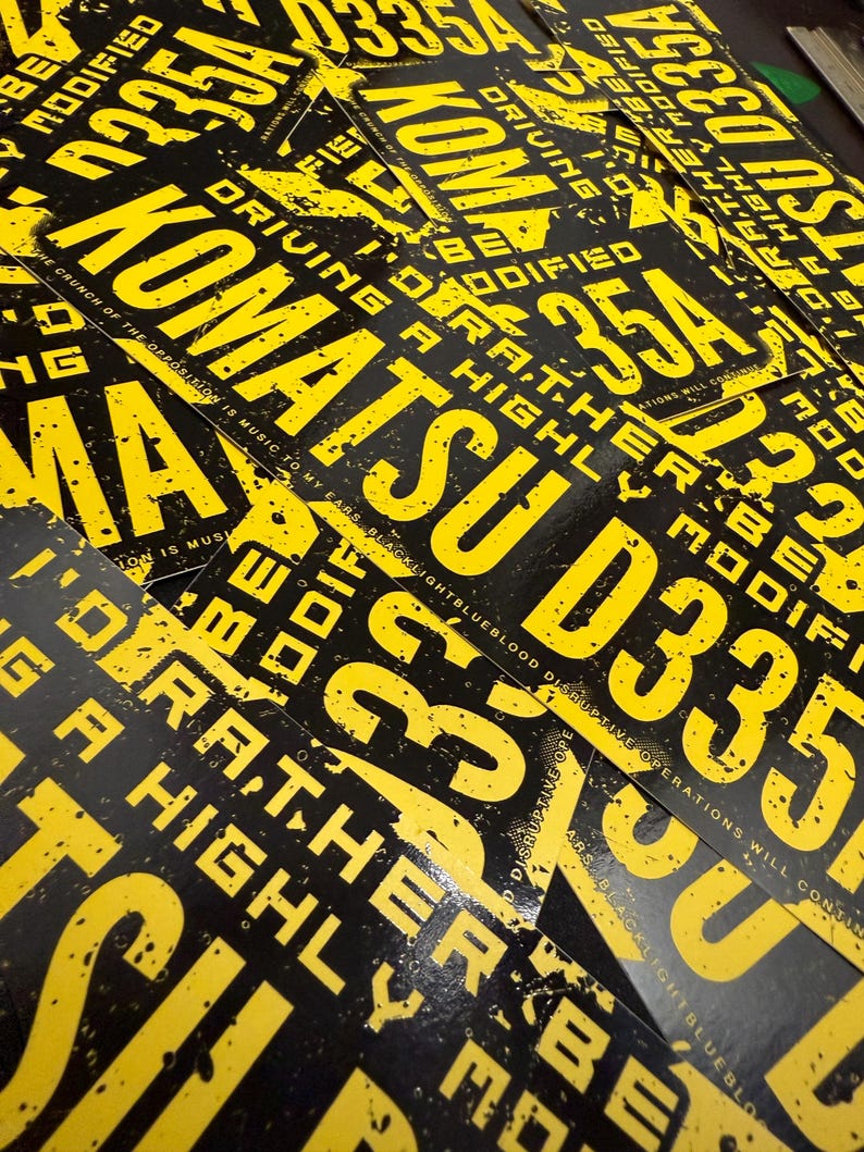 Puede incluir: Una colecci&oacute;n de impresiones gr&aacute;ficas en negro y amarillo. Las impresiones presentan las palabras "KOMATSU D335A" en grandes letras amarillas, junto con otro texto. El dise&ntilde;o general tiene una est&eacute;tica grunge.