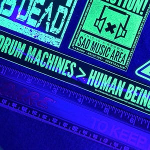May include: A close-up of a ruler with a green and black sticker that reads "Drum Machines > Human Beings". The sticker is on a black background and has a distressed, vintage look. The ruler is marked in inches.