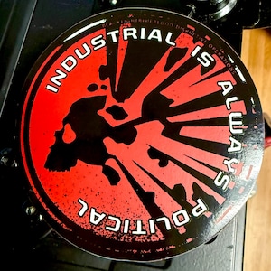 May include: A round sticker with a black background and red accents. The sticker features a skull graphic and the words "INDUSTRIAL IS ALWAYS POLITICAL" in white. The design has a distressed, grunge aesthetic.