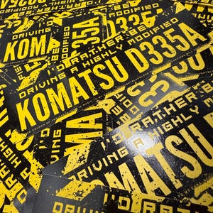 Puede incluir: Una colecci&oacute;n de pegatinas negras y amarillas. Las pegatinas presentan el texto "KOMATSU D335A" y "DRIVING A HIGHLY MODIFIED" en una fuente audaz y desgastada. El dise&ntilde;o tiene una est&eacute;tica grunge.