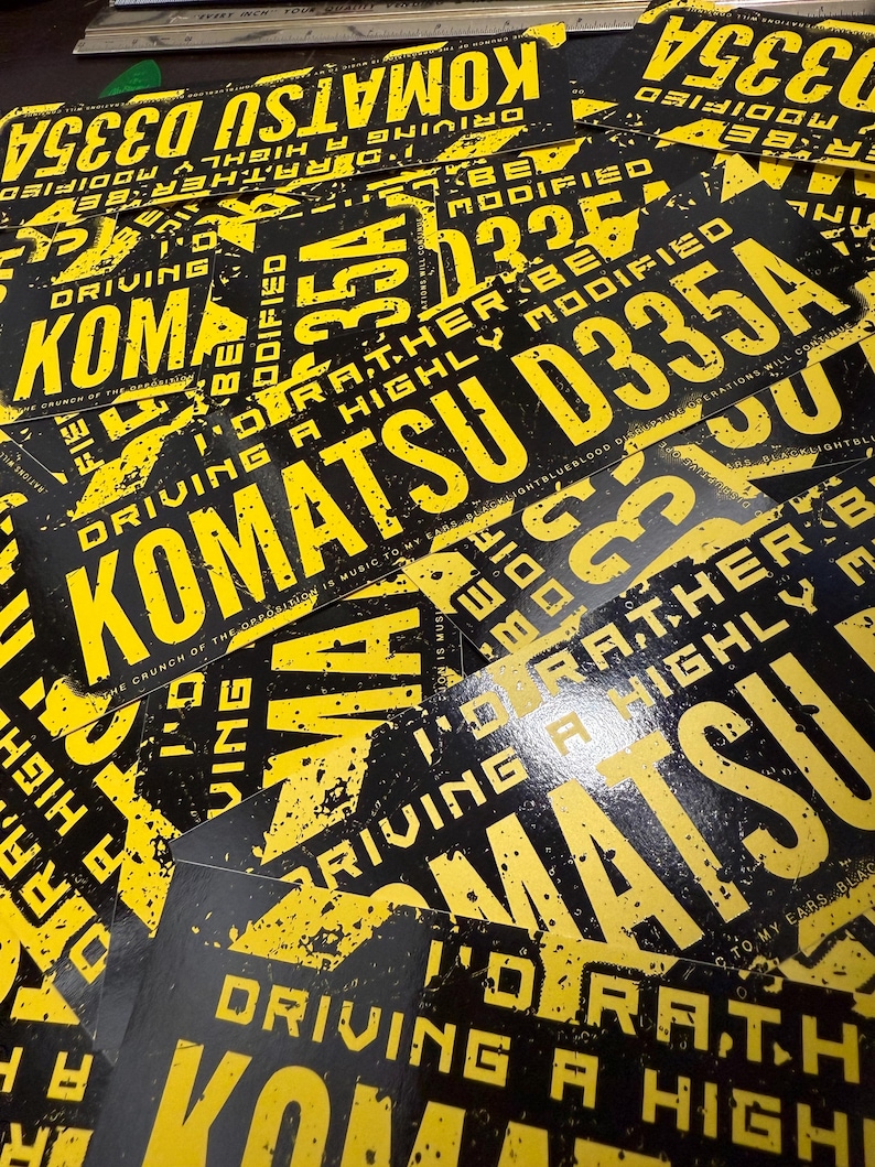 Puede incluir: Una colecci&oacute;n de pegatinas negras y amarillas. Las pegatinas presentan el texto "KOMATSU D335A" y "DRIVING A HIGHLY MODIFIED" en una fuente audaz y desgastada. El dise&ntilde;o tiene una est&eacute;tica grunge.