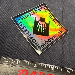 May include: A black and white sticker with a holographic finish. The sticker features a hand with a skeleton hand inside a plastic bag. The text on the sticker reads "Shopelessly Seduced by the Music of the Shadows".