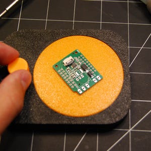 May include: A green circuit board with various electronic components is placed on an orange circular pad. A person's finger is holding a small orange disc. Soldering tools and tweezers are also visible.