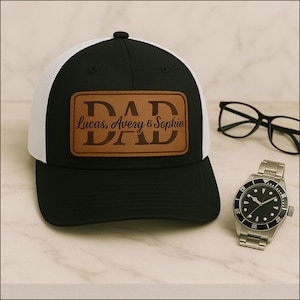 Op de afbeelding: Zwarte en witte truckerpet met een bruin leren patch met de tekst "DAD" en de namen "Lucas, Avery & Sophia". Een zilveren horloge en een zwarte bril zijn ook zichtbaar.