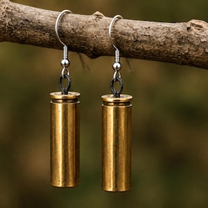 May include: A pair of drop earrings featuring cylindrical brass bullet casings as the primary pendants. Each casing is attached to a silver-toned metal fishhook ear wire by a small black jump ring, creating a dangling design with a metallic finish.