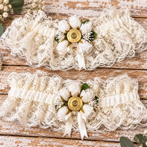 May include: Two ivory lace garters featuring a central cluster of white fabric roses and green leaves. Each garter is accented with a gold-toned Remington 12 GA shotgun shell casing base and a small white satin ribbon bow at the center.