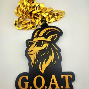 Peut inclure: Un collier en chaîne doré est placé au-dessus d'un pendentif noir avec un motif de tête de chèvre dorée portant des lunettes de soleil. Le pendentif porte les lettres dorées "G.O.A.T." en bas.