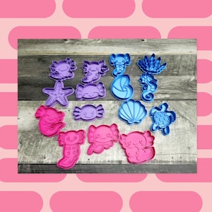 May include: A collection of colorful sea creature-shaped cookie cutters. The set includes axolotls, starfish, crabs, seahorses, seashells, and turtles in shades of purple, blue, and pink. These plastic cutters are ideal for making themed cookies.