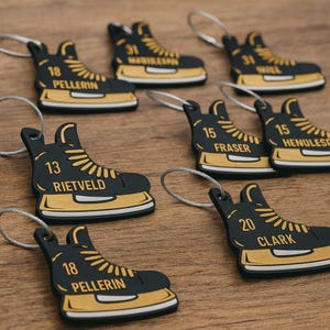 May include: A collection of black and gold hockey skate keychains with various numbers and names printed on them. The keychains are arranged on a wooden surface. The text includes names like "Pellerin", "Rietveld", "Fraser", and "Clark".