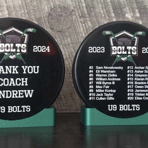May include: Two black hockey pucks with green bases. The puck on the left has the text "2023 BOLTS 2024" and "THANK YOU COACH ANDREW U9 BOLTS". The puck on the right has the text "2023 BOLTS 2024" and a list of player names and numbers.