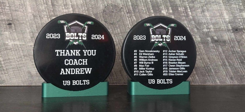 May include: Two black hockey pucks with green bases. The puck on the left has the text "2023 BOLTS 2024" and "THANK YOU COACH ANDREW U9 BOLTS". The puck on the right has the text "2023 BOLTS 2024" and a list of player names and numbers.