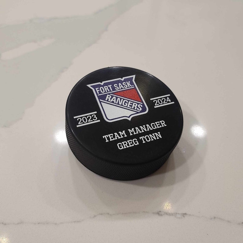 May include: Black hockey puck with a white and blue logo that says "Fort Sask. Rangers" and "Team Manager Greg Tonn". The puck also has the years "2023" and "2024" printed on it.