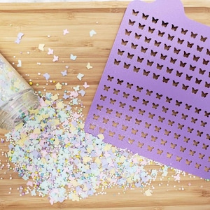 Puede incluir: Una plantilla de plástico púrpura con una cuadrícula de formas de mariposa. La plantilla está sobre una superficie de madera con una dispersión de espolvoreados de colores pastel.