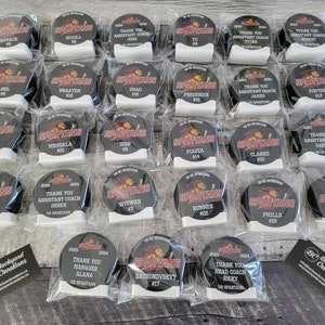 May include: A collection of 20 black and red hockey puck-shaped awards with white lettering. The awards are individually packaged in clear plastic bags. The awards feature the words "Thank You" and "Assistant Coach" along with the names of the recipients. The awards also feature the words "Thank You Manager" and "Thank You Head Coach" along with the names of the recipients. The awards also feature the words "Spartans" and a red Spartan logo.