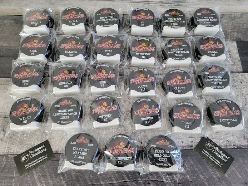 May include: A collection of 20 black and red hockey puck-shaped awards with white lettering. The awards are individually packaged in clear plastic bags. The awards feature the words "Thank You" and "Assistant Coach" along with the names of the recipients. The awards also feature the words "Thank You Manager" and "Thank You Head Coach" along with the names of the recipients. The awards also feature the words "Spartans" and a red Spartan logo.