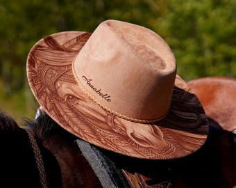 May include: A tan cowboy hat with a detailed horse head design and the name "Annabelle" is resting on a saddle. The hat has a rounded crown and a wide brim with a decorative band.