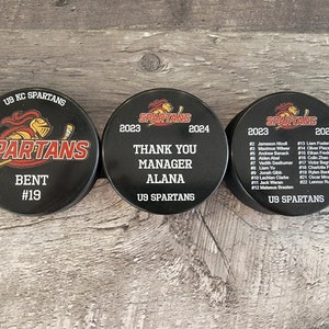 May include: Three black hockey pucks with the team name "SPARTANS" printed on them. The first puck has the player's name "BENT" and number "#19". The second puck says "THANK YOU MANAGER ALANA" and the third puck has a list of player names and numbers.
