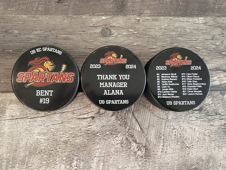 May include: Three black hockey pucks with the team name "SPARTANS" printed on them. The first puck has the player's name "BENT" and number "#19". The second puck says "THANK YOU MANAGER ALANA" and the third puck has a list of player names and numbers.