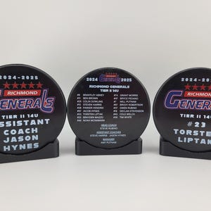 May include: Three black hockey pucks with white text and logos. The left puck says "2024-2025 Richmond Generals Tier II 14U Assistant Coach Jason Hynes". The middle puck has a roster of players and coaches for the Richmond Generals Tier II 14U team. The right puck says "2024-2025 Richmond Generals Tier II 14U #23 Torsten Liptak".