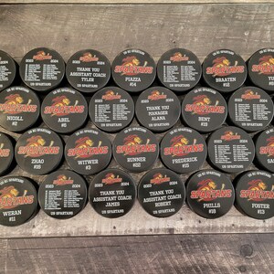 May include: A collection of 20 black and red hockey pucks with the word "SPARTANS" and a number on each puck. The pucks are arranged in a circle on a wooden surface.