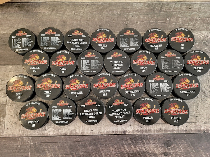 May include: A collection of 20 black and red hockey pucks with the word "SPARTANS" and a number on each puck. The pucks are arranged in a circle on a wooden surface.