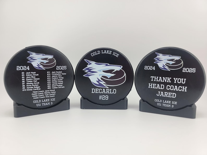 May include: Three black hockey pucks with white and blue designs. The center puck says "Thank You Head Coach Jared" and "Cold Lake Ice U11 Team 3". The left puck has a list of player names and numbers for the 2024-2025 season. The right puck says "Decarlo #29" and "Cold Lake Ice".