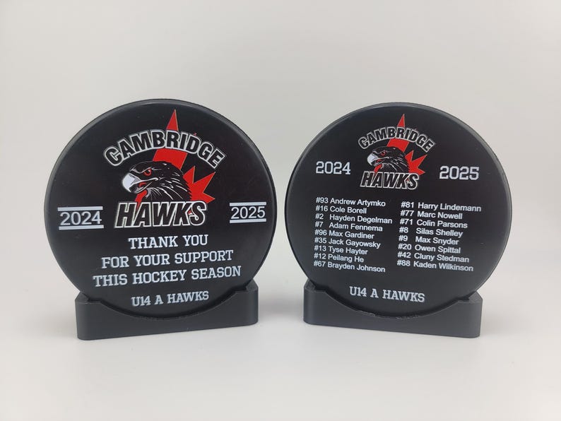 May include: Two black hockey pucks with a white base. The pucks have a black and red logo with the text "Cambridge Hawks" and the years "2024" and "2025". The pucks also have a list of player names and numbers. The text on the pucks reads "Thank you for your support this hockey season" and "U14 A Hawks".