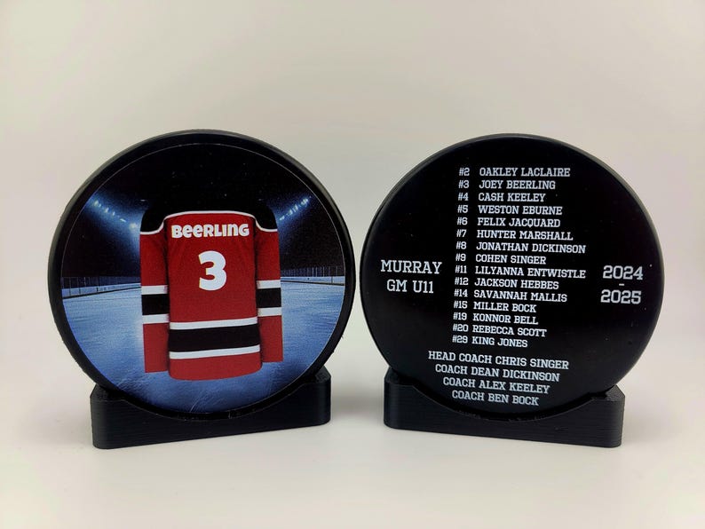 May include: Black hockey puck with a white hockey jersey with the name "Beerling" and the number "3" on the back. The puck is on a black stand. The other side of the puck has the text "Murray GM U11" and a list of names and numbers. The text "2024" and "2025" are also on the puck.