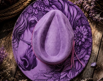 May include: A purple felt hat featuring a detailed black ink illustration of a horse head surrounded by floral patterns on the wide brim. The crown is teardrop-shaped and encircled by a thin, matching purple cord with a small tassel detail.
