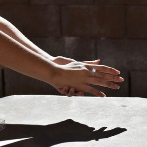 May include: A close-up of a hand wearing a ring with a light blue gemstone. The hand is resting on a light gray surface.