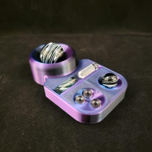May include: A purple and blue glass marble holder with a compartment for a small glass tube. The holder has a round top with a black and white marble inside. There are three clear marbles in the bottom compartment.