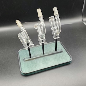 May include: Three clear glass smoking accessories with angled stems and flared tops are displayed on a green rectangular base with black stems. The glass pieces have a curved design. The background is a dark green.