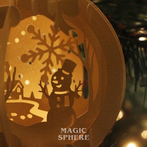 May include: A brown paper mache Christmas ornament with a cut-out design of a snowman, a snowflake, and a winter scene. The ornament is lit from inside, creating a warm glow. The text "MAGIC SPHERE" is printed on the bottom of the ornament.