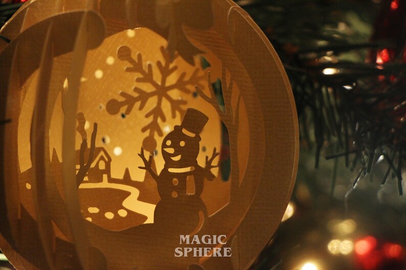 May include: A brown paper mache Christmas ornament with a cut-out design of a snowman, a snowflake, and a winter scene. The ornament is lit from inside, creating a warm glow. The text "MAGIC SPHERE" is printed on the bottom of the ornament.