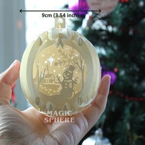 May include: A white, 3.54 inch, paper Christmas ornament with a winter scene inside. The scene includes a snowman, a house, and a star. The ornament is labeled "MAGIC SPHERE".