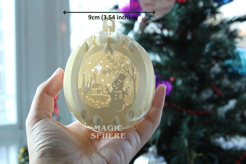 May include: A white, 3.54 inch, paper Christmas ornament with a winter scene inside. The scene includes a snowman, a house, and a star. The ornament is labeled "MAGIC SPHERE".