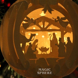 May include: A paper mache Christmas ornament with a cut-out design depicting the nativity scene. The ornament is illuminated from within, casting a warm glow on the figures. The text "MAGIC SPHERE" is printed on the bottom of the ornament.