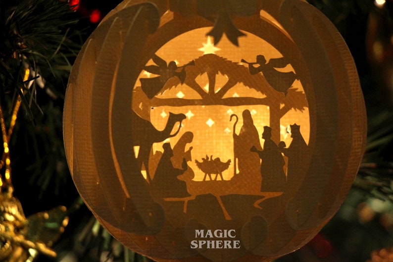 May include: A paper mache Christmas ornament with a cut-out design depicting the nativity scene. The ornament is illuminated from within, casting a warm glow on the figures. The text "MAGIC SPHERE" is printed on the bottom of the ornament.