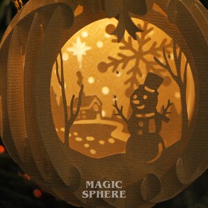May include: A paper Christmas ornament with a cut-out design of a snowman, a house, and a snowflake. The ornament is illuminated from within, creating a warm glow. The text "MAGIC SPHERE" is printed on the bottom of the ornament.