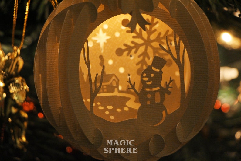 May include: A paper Christmas ornament with a cut-out design of a snowman, a house, and a snowflake. The ornament is illuminated from within, creating a warm glow. The text "MAGIC SPHERE" is printed on the bottom of the ornament.