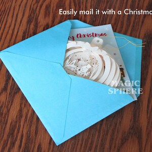 May include: A blue envelope with a white Christmas card and a white paper ornament inside. The ornament has a snowflake design and the text "Magic Sphere" on it. The card has the text "Merry Christmas" on it. Easily mail it with a Christmas card!