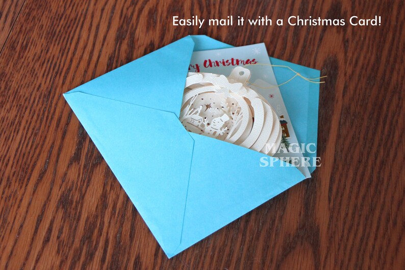 May include: A blue envelope with a white Christmas card and a white paper ornament inside. The ornament has a snowflake design and the text "Magic Sphere" on it. The card has the text "Merry Christmas" on it. Easily mail it with a Christmas card!