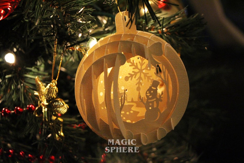 May include: A white paper Christmas ornament with a snowman and snowflake design. The ornament is a 3D paper sphere with the words "Magic Sphere" printed on the bottom.