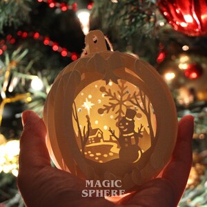 May include: A white paper Christmas ornament with a cut-out design of a winter scene. The ornament is lit from inside, creating a warm glow. The scene includes a snowman, a house, a tree, and a star. The ornament is labeled "MAGIC SPHERE".