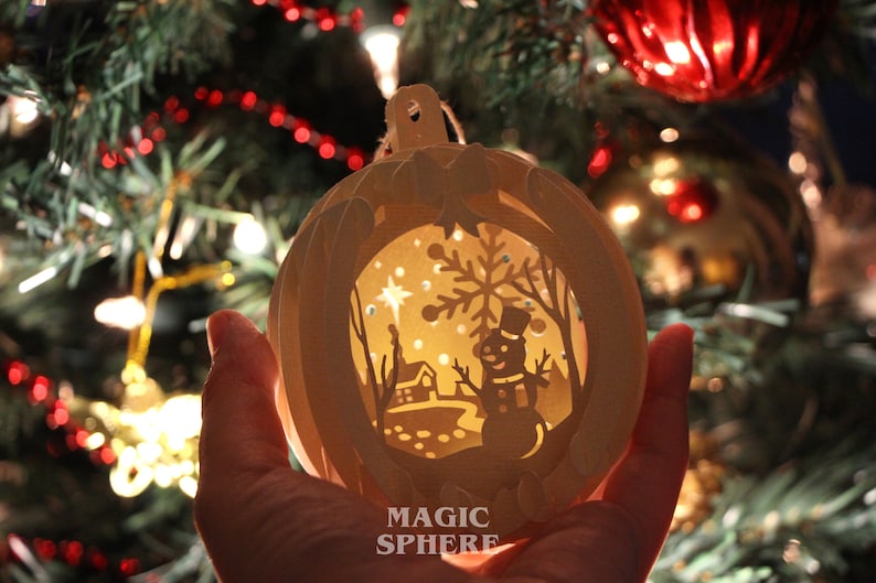 May include: A white paper Christmas ornament with a cut-out design of a winter scene. The ornament is lit from inside, creating a warm glow. The scene includes a snowman, a house, a tree, and a star. The ornament is labeled "MAGIC SPHERE".