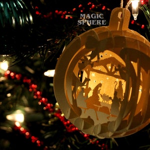 May include: A white paper ornament shaped like a sphere with a cut-out design of a nativity scene. The ornament is hanging on a Christmas tree with red and white lights. The text "MAGIC SPHERE" is printed on the ornament.