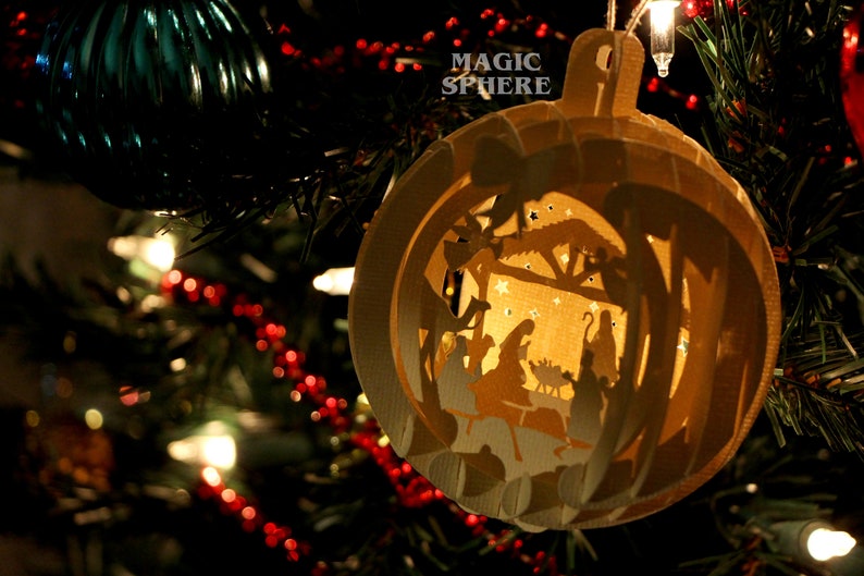 May include: A white paper ornament shaped like a sphere with a cut-out design of a nativity scene. The ornament is hanging on a Christmas tree with red and white lights. The text "MAGIC SPHERE" is printed on the ornament.
