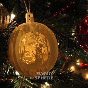 May include: A white, paper Christmas ornament with a cut-out design of a snowman, a house, and a star. The ornament is lit from inside, creating a warm glow. The text "MAGIC SPHERE" is printed on the bottom of the ornament.